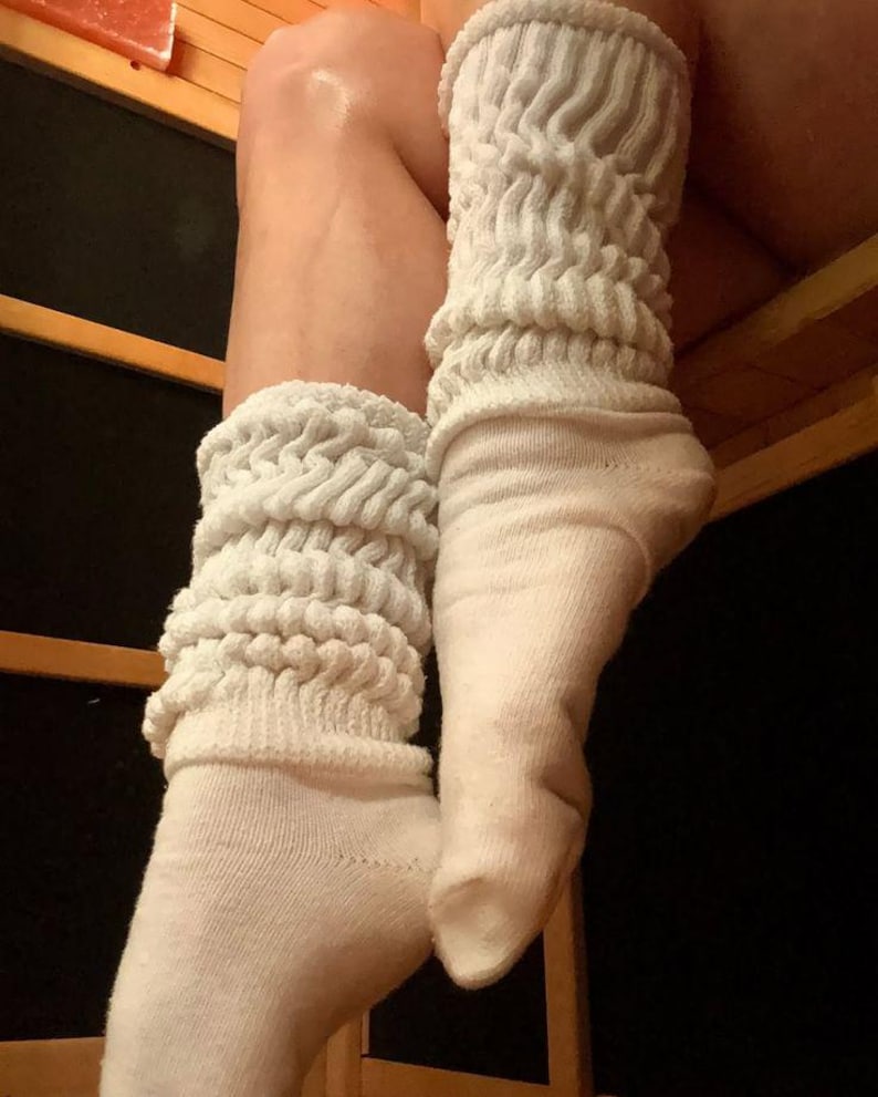 May include: A pair of off-white slouch socks. The top portion of the socks is a thick, textured knit, while the foot portion is a smooth knit. The socks are pulled up, creating a scrunched effect. The background is a wooden structure.