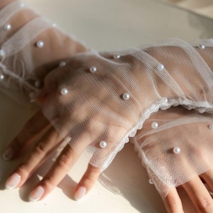 Puede incluir: Guantes blancos y transparentes sin dedos adornados con perlas redondas y dispersas. Los guantes tienen un delicado ribete de encaje. La imagen muestra la textura y la colocaci&oacute;n de las perlas en los guantes.