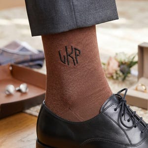 Peut inclure: Une chaussette mi-mollet marron ornée d'un monogramme noir brodé avec les lettres UKP. La chaussette est portée avec une chaussure richelieu en cuir noir et un pantalon gris foncé, présentant une texture en maille lisse et un design ajusté.