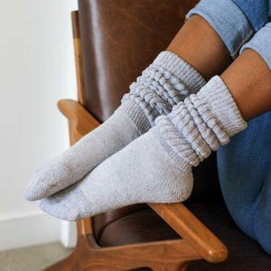 May include: A pair of light gray, slouchy knit socks. The socks are pulled up, with a ribbed cuff and a textured, folded design. The socks are worn with blue jeans, and the person is sitting in a wooden chair.