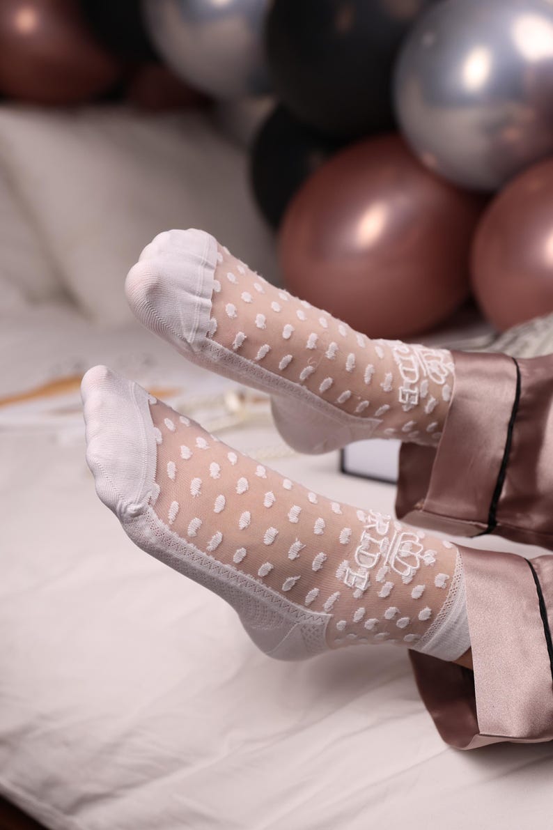 May include: White sheer socks with a polka dot pattern and floral lace detailing. The socks are worn with a satin pajama set. The background includes balloons in shades of silver, black, and rose gold.