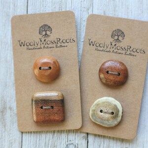 May include: Four buttons attached to two brown rectangular cards labeled Wooly Moss Roots Handmade Artisan Buttons. The buttons include a smooth round light brown one, a square brown one, a round dark brown one, and a round off-white one.