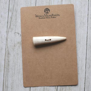 May include: A light-colored, wooden toggle button with two holes, displayed on a brown card. The card has the text "Wooly Moss Roots Handmade Artisan Buttons" and a tree logo. The background is a light-colored, wooden surface.