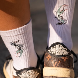 Peut inclure: Chaussettes de sport blanches avec un motif de carpe koï brodé. Les chaussettes sont associées à des baskets marron et blanches avec le logo Air Jordan et un motif.