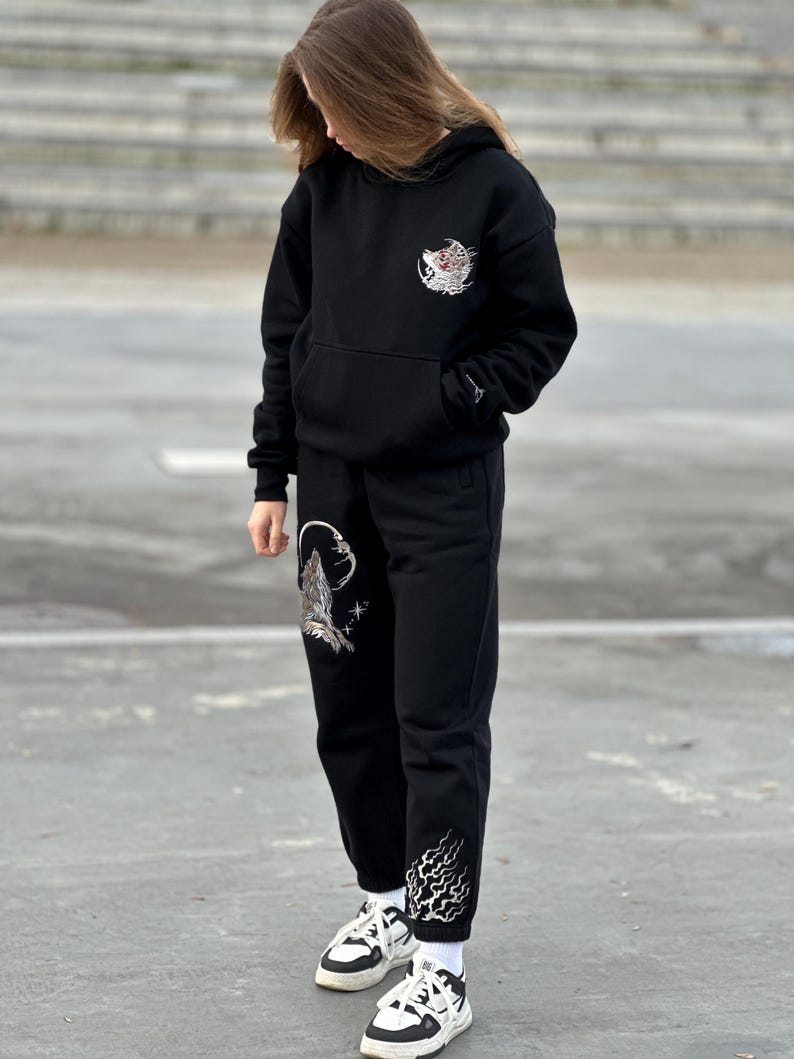 May include: Black hoodie and sweatpants set with embroidered designs. The hoodie features a design on the chest, and the sweatpants have a design on the left leg and ankle. The set is paired with white and black sneakers.
