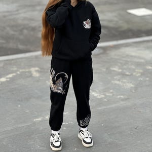 May include: Black hoodie and sweatpants set with wolf graphics. The hoodie has a front pocket and a small wolf design on the chest. The sweatpants feature a large wolf howling at the moon design on the left leg.