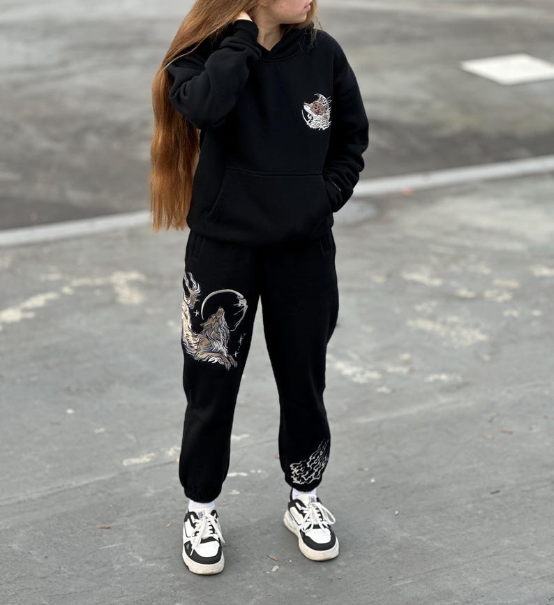 May include: Black hoodie and sweatpants set with wolf graphics. The hoodie has a front pocket and a small wolf design on the chest. The sweatpants feature a large wolf howling at the moon design on the left leg.