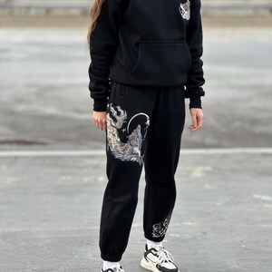 May include: Black hoodie and sweatpants set with a wolf graphic. The hoodie has a front pocket and a small graphic on the chest. The sweatpants feature a large wolf design on the left leg and a smaller design on the right leg.