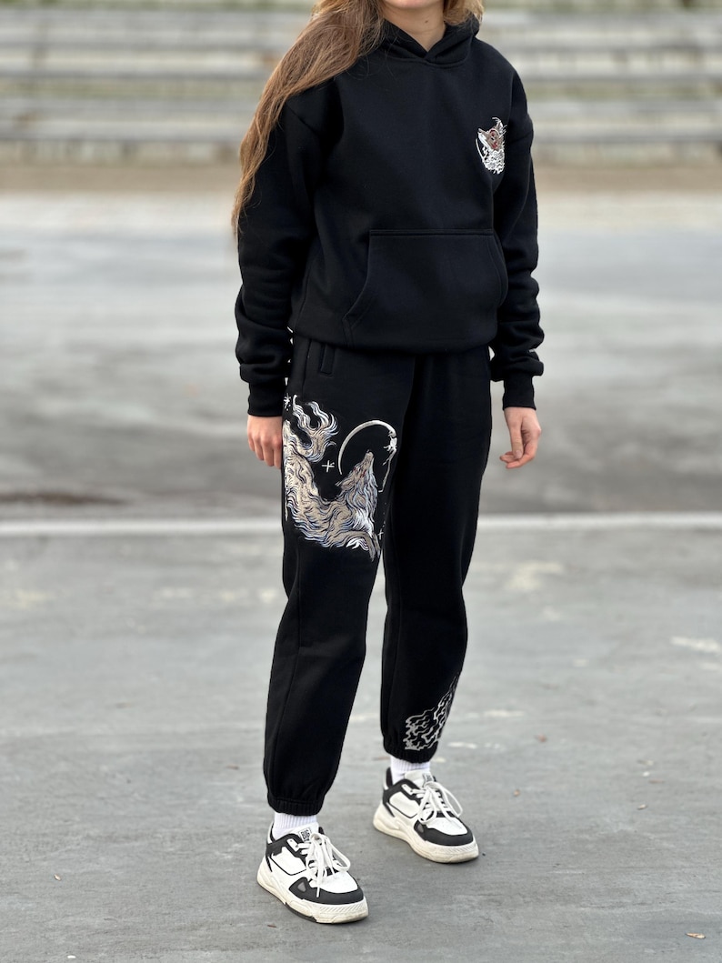May include: Black hoodie and sweatpants set with a wolf graphic. The hoodie has a front pocket and a small graphic on the chest. The sweatpants feature a large wolf design on the left leg and a smaller design on the right leg.