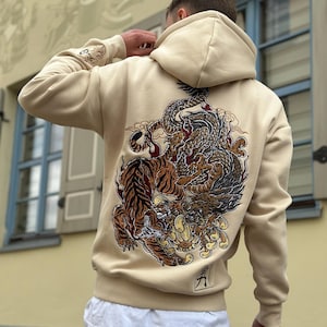 May include: Beige hoodie featuring a detailed graphic of a tiger and dragon in shades of brown, red, and black on the back. The hoodie has a drawstring hood and a small logo near the bottom. The sleeves have a small design on the upper arm.