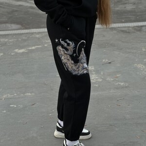 May include: Black sweatpants with a detailed wolf graphic on the left leg, featuring a wolf howling at the moon and stars. The pants are paired with black and white sneakers and a black hoodie.