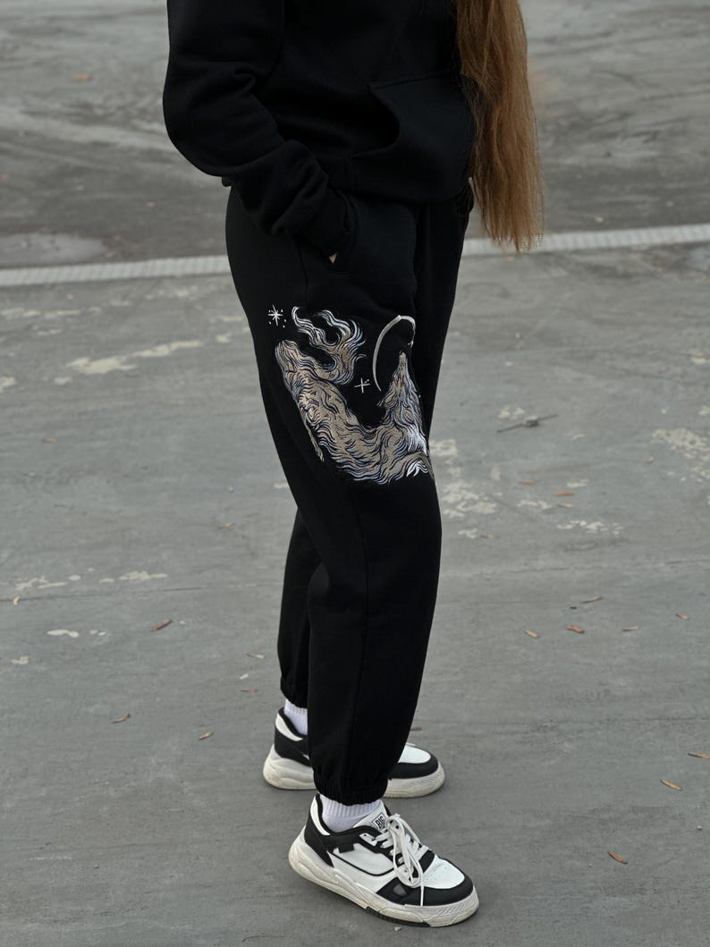 May include: Black sweatpants with a detailed wolf graphic on the left leg, featuring a wolf howling at the moon and stars. The pants are paired with black and white sneakers and a black hoodie.
