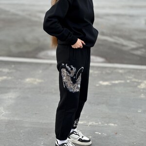 May include: Black hoodie and sweatpants set. The sweatpants feature a detailed silver and white graphic design on the left leg. The outfit is paired with white and black sneakers.