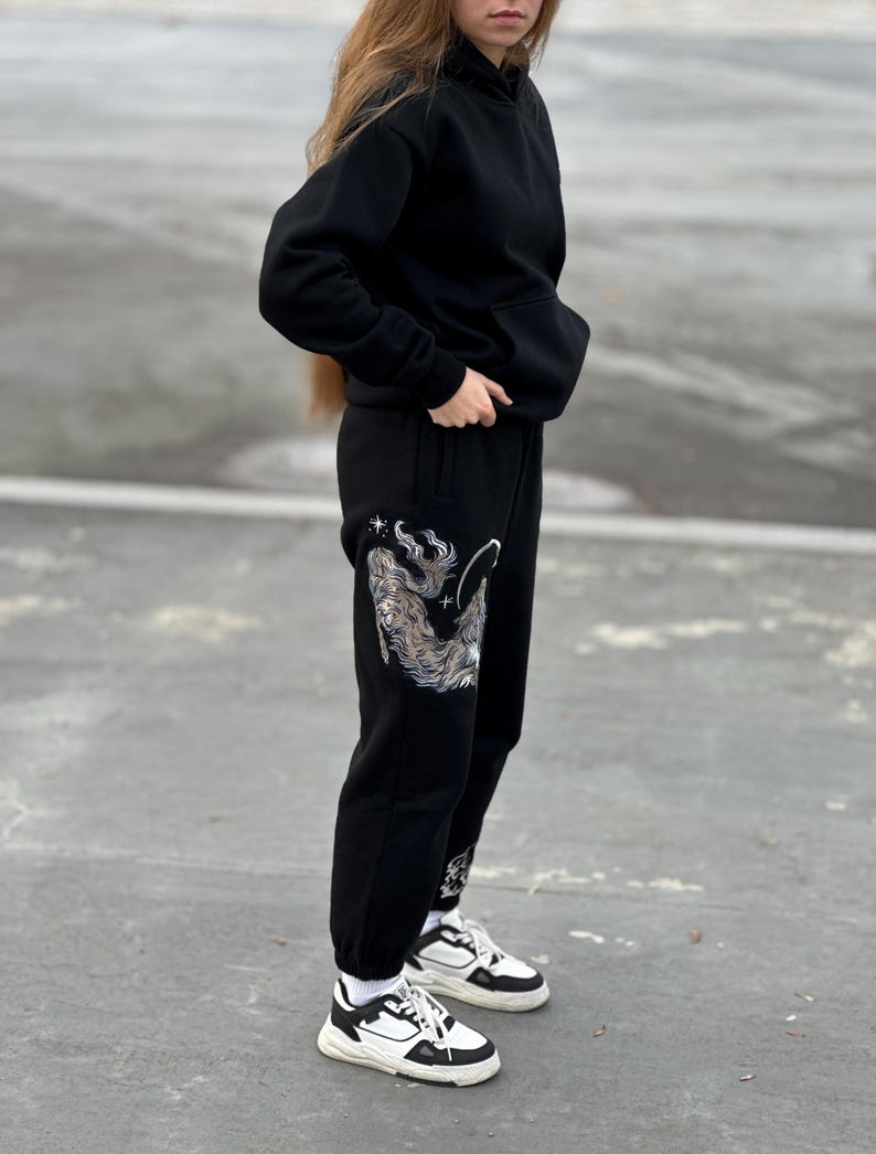 May include: Black hoodie and sweatpants set. The sweatpants feature a detailed silver and white graphic design on the left leg. The outfit is paired with white and black sneakers.