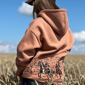 Op de afbeelding: Een stoffig roze hoodie met een western-thema grafiek op de rug, met ruiters te paard tegen een woestijnlandschap. De hoodie heeft een capuchon en wordt gecombineerd met een crèmekleurige baseballcap met een bruin vizier en een paardenontwerp.