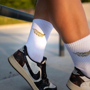 Könnte beinhalten: Weiße gerippte Socken mit einem goldenen Blattdesign, getragen mit schwarzen, weißen und braunen Turnschuhen. Die Sneaker haben ein einzigartiges Muster und einen schwarzen Nike-Swoosh. Die Schuhe befinden sich auf einer Betonoberfläche.