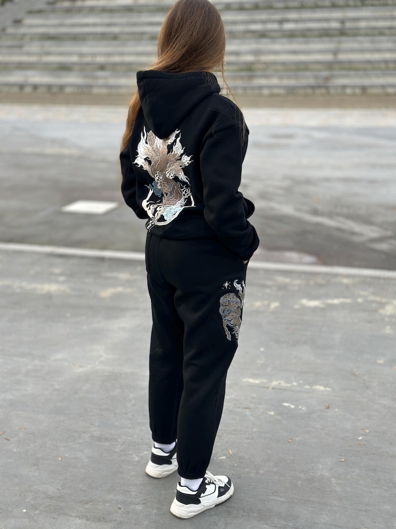 May include: Black hoodie and sweatpants set with embroidered dragon and tiger designs. The hoodie features a large dragon design on the back, while the sweatpants have a tiger design on the leg. The set is worn with white sneakers.