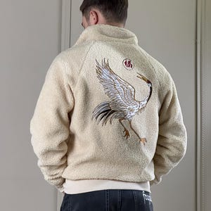 Puede incluir: Una chaqueta de forro polar de color crema con un gran diseño de grúa bordado en la espalda. La grúa es de color dorado y negro con un círculo rojo que contiene el carácter chino "福" (fú) que significa "buena fortuna" o "felicidad".