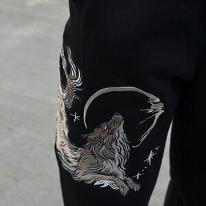 May include: Black sweatpants with an embroidered design of a howling wolf under a crescent moon. The wolf is depicted in shades of brown, gray, and white, with red accents. The design includes stars and a stylized cloud.