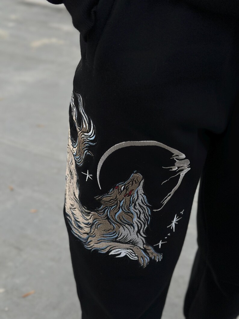 May include: Black sweatpants with an embroidered design of a howling wolf under a crescent moon. The wolf is depicted in shades of brown, gray, and white, with red accents. The design includes stars and a stylized cloud.