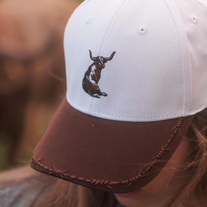 May include: A white baseball cap with a brown brim and a brown button on top. The front of the cap features an embroidered brown and white longhorn steer graphic. The brown brim is decorated with an embroidered black barbed wire pattern.