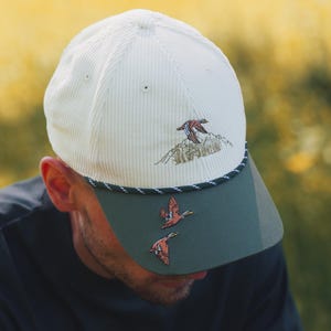May include: Cream corduroy baseball cap with a green peak and embroidered flying ducks. The cap has a black and white rope detail and a mountain scene with a duck. A stylish accessory for outdoor pursuits.