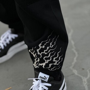 May include: Black sweatpants with a white flame design on the lower leg. The pants are paired with black and white Puma sneakers with white laces. The Puma logo is visible on the tongue and side of the shoe.