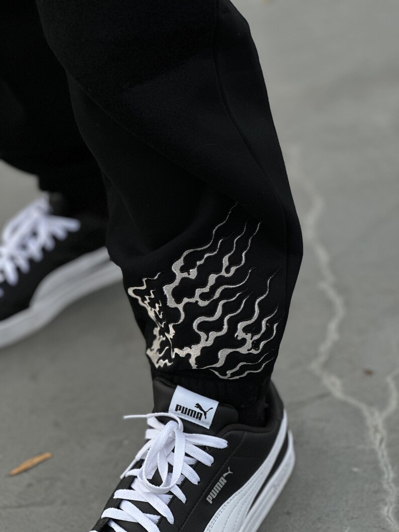 May include: Black sweatpants with a white flame design on the lower leg. The pants are paired with black and white Puma sneakers with white laces. The Puma logo is visible on the tongue and side of the shoe.