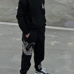 May include: Black hoodie and sweatpants set with a detailed wolf design on the left leg and upper chest. The hoodie has a front pocket and a hood. The sweatpants have a drawstring waist and cuffed ankles. The outfit is paired with black and white sneakers.