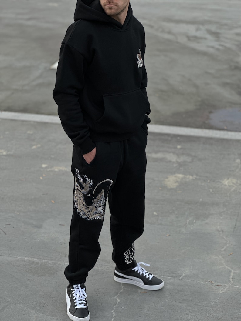 May include: Black hoodie and sweatpants set with a detailed wolf design on the left leg and upper chest. The hoodie has a front pocket and a hood. The sweatpants have a drawstring waist and cuffed ankles. The outfit is paired with black and white sneakers.