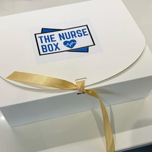 Op de afbeelding: Een witte geschenkdoos met een gebogen deksel, vastgebonden met een gouden lint. De doos heeft een sticker met de tekst "THE NURSE BOX" in blauw en wit, met een hartsymbool.