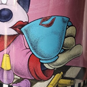 May include: A pink ceramic mug featuring a cartoon character with red lips, holding a blue teacup with red lipstick marks. The mug has a black and white checkered base and illustrations of makeup.