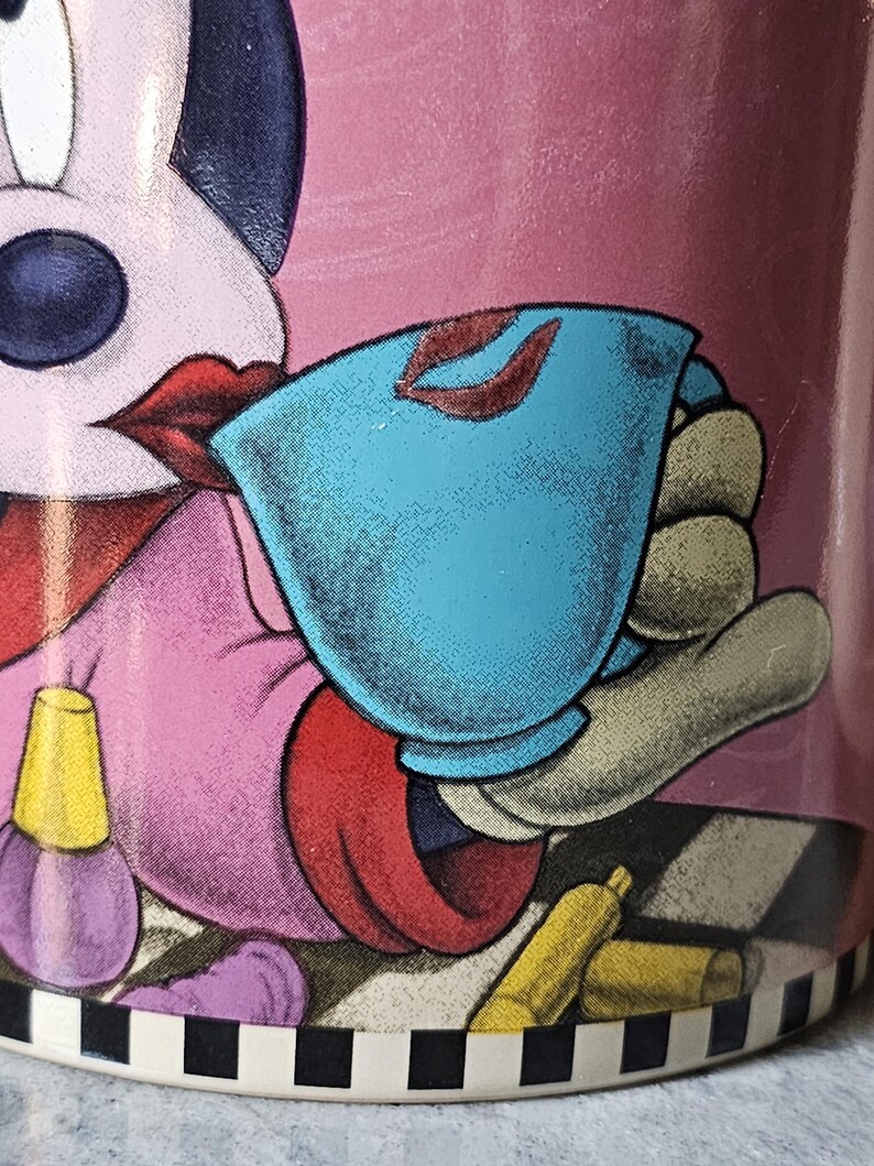 May include: A pink ceramic mug featuring a cartoon character with red lips, holding a blue teacup with red lipstick marks. The mug has a black and white checkered base and illustrations of makeup.