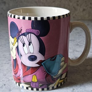 May include: Pink ceramic mug featuring Minnie Mouse in a stylish outfit. The mug has a cream-colored interior and a tan handle. The design includes a black and white checkered pattern at the top and bottom.