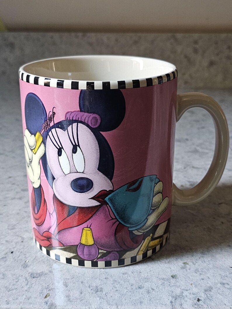 May include: Pink ceramic mug featuring Minnie Mouse in a stylish outfit. The mug has a cream-colored interior and a tan handle. The design includes a black and white checkered pattern at the top and bottom.