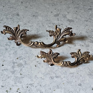 May include: Two ornate, antique-style bronze drawer pulls with a curved handle and decorative leaf design. The pulls are set against a textured, gray background, showcasing their intricate details and aged finish. These pulls are ideal for vintage furniture restoration.