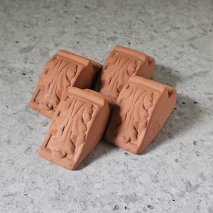 May include: A set of four terracotta-colored clay pot feet, each featuring a molded acanthus leaf design. These small, wedge-shaped supports have a flat top surface and a textured, carved front panel, designed to elevate containers off the ground.