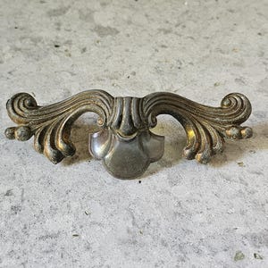 May include: An antique bronze-toned drawer pull with ornate, symmetrical scrollwork. The central portion features a shield-like design, and the overall aesthetic suggests a vintage or Victorian style. The hardware is likely made of metal.