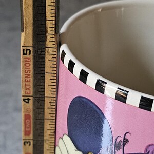 May include: A pink ceramic mug featuring a cartoon character design. The mug has a black and white striped rim and a light-colored interior. A wooden ruler is placed next to the mug, showing a measurement of approximately 5 inches.