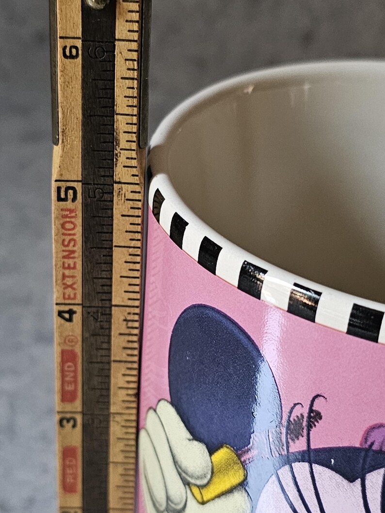 May include: A pink ceramic mug featuring a cartoon character design. The mug has a black and white striped rim and a light-colored interior. A wooden ruler is placed next to the mug, showing a measurement of approximately 5 inches.