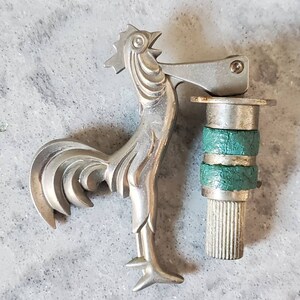 Puede incluir: Un encendedor de plata con forma de gallo con una base verde y plateada. El encendedor tiene una palanca articulada en el lateral y una base texturizada.
