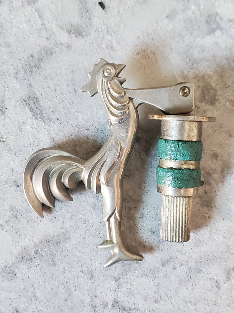 Puede incluir: Un encendedor de plata con forma de gallo con una base verde y plateada. El encendedor tiene una palanca articulada en el lateral y una base texturizada.
