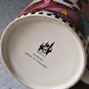 May include: A ceramic mug featuring Minnie Mouse in a pink dress. The mug has a black and white checkered pattern around the rim. The bottom of the mug has the Disney logo and text that reads "Made in Thailand."