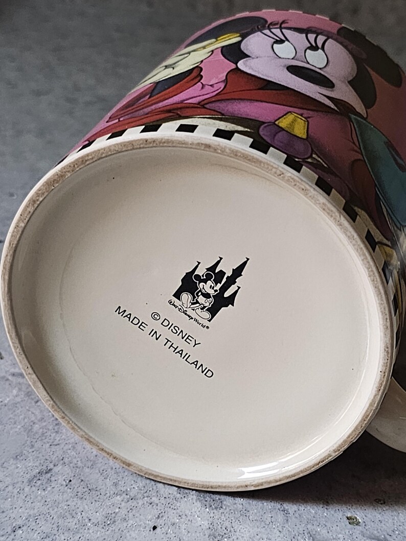 May include: A ceramic mug featuring Minnie Mouse in a pink dress. The mug has a black and white checkered pattern around the rim. The bottom of the mug has the Disney logo and text that reads "Made in Thailand."