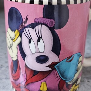 May include: Pink ceramic mug featuring Minnie Mouse applying makeup. The mug has a black and white striped rim and base. Minnie Mouse is depicted in a pink robe, holding a blue compact and a yellow bottle. The mug has a white interior.