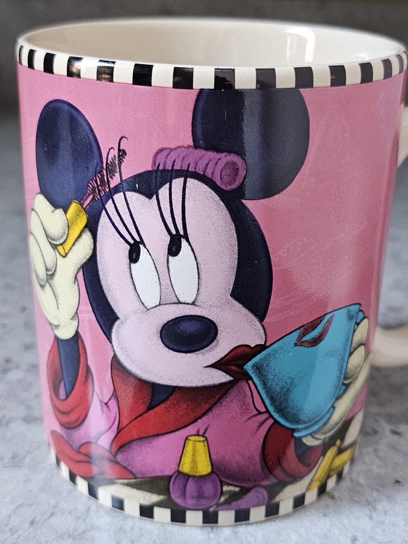 May include: Pink ceramic mug featuring Minnie Mouse applying makeup. The mug has a black and white striped rim and base. Minnie Mouse is depicted in a pink robe, holding a blue compact and a yellow bottle. The mug has a white interior.