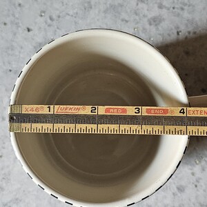 May include: Cream-colored mug with a black dashed border. A wooden ruler is placed inside the mug, measuring approximately 3.5 inches across. The mug has a simple, classic design.