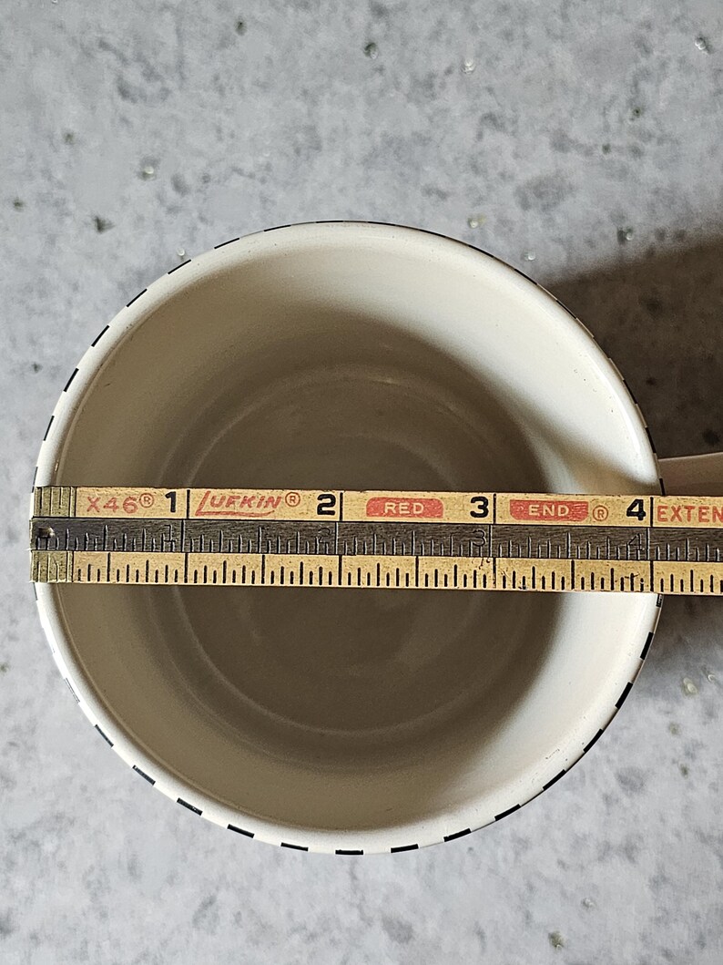 May include: Cream-colored mug with a black dashed border. A wooden ruler is placed inside the mug, measuring approximately 3.5 inches across. The mug has a simple, classic design.