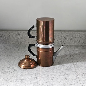 May include: A two-tiered copper coffee maker with black handles and a silver-toned spout. The cylindrical device features a removable copper lid with a knob handle, a metallic silver rim connecting the two sections, and a polished copper finish.