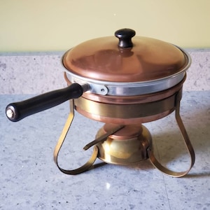 May include: A copper chafing dish with a black handle and a brass stand. The dish has a lid with a black knob.